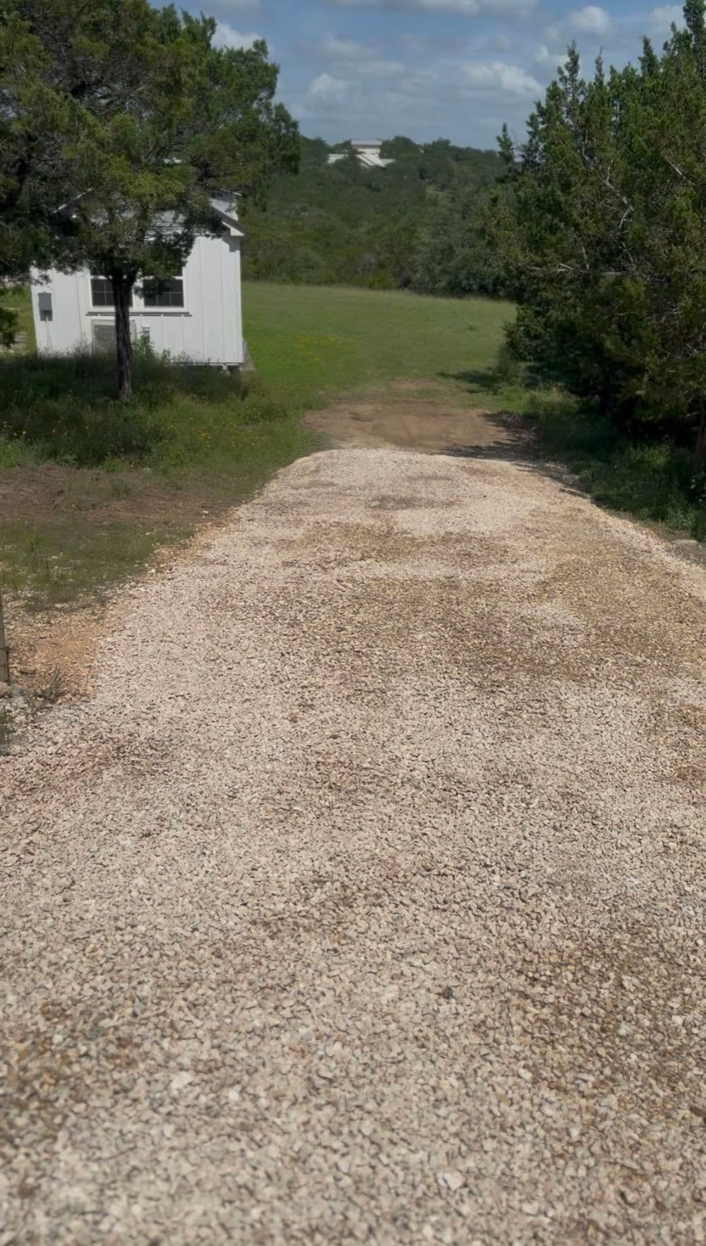 Gravel driveway leading to a white building with green landscape and trees nearby.