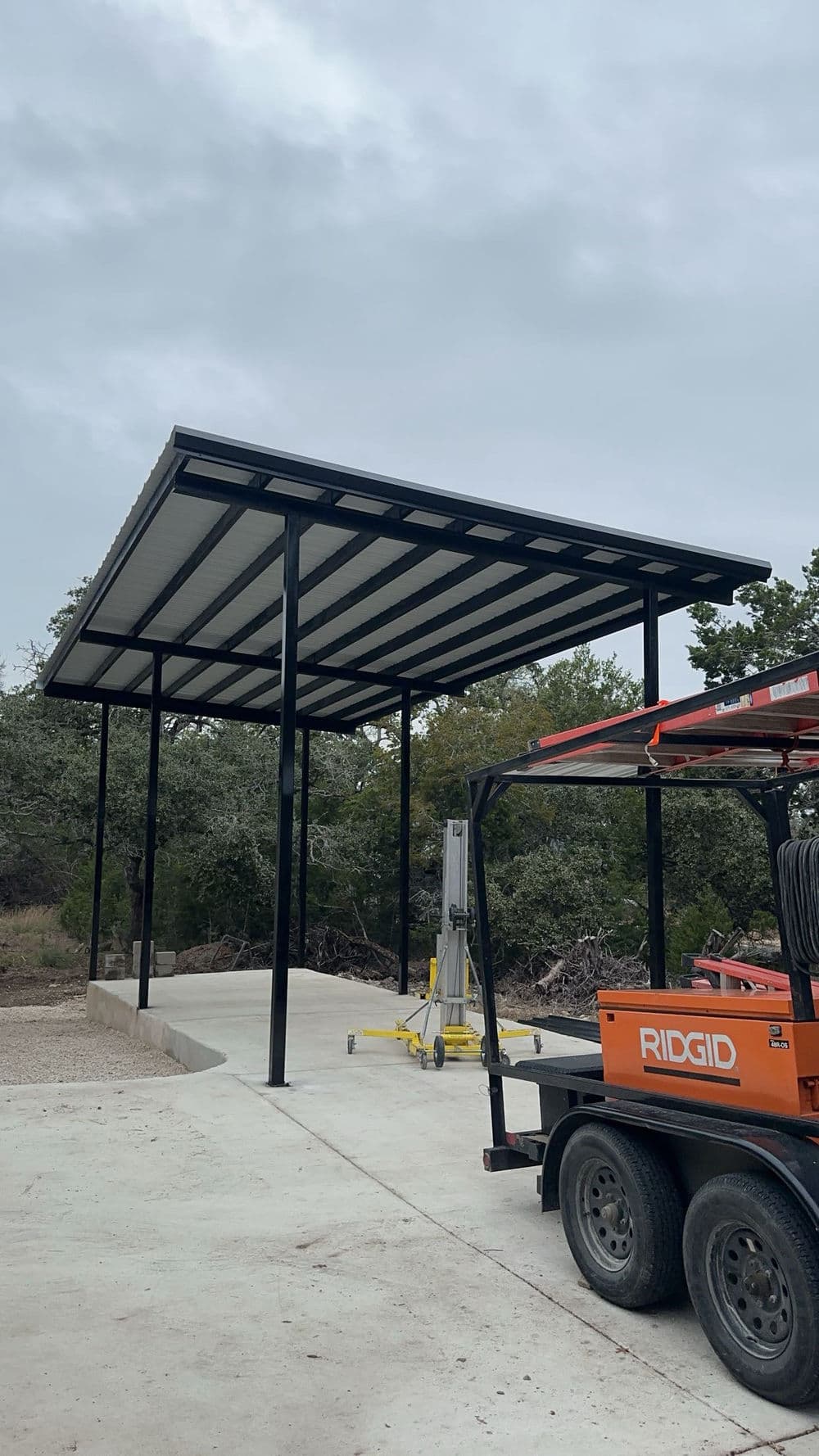 Steel carport structure under construction in a rural setting with a trailer nearby.
