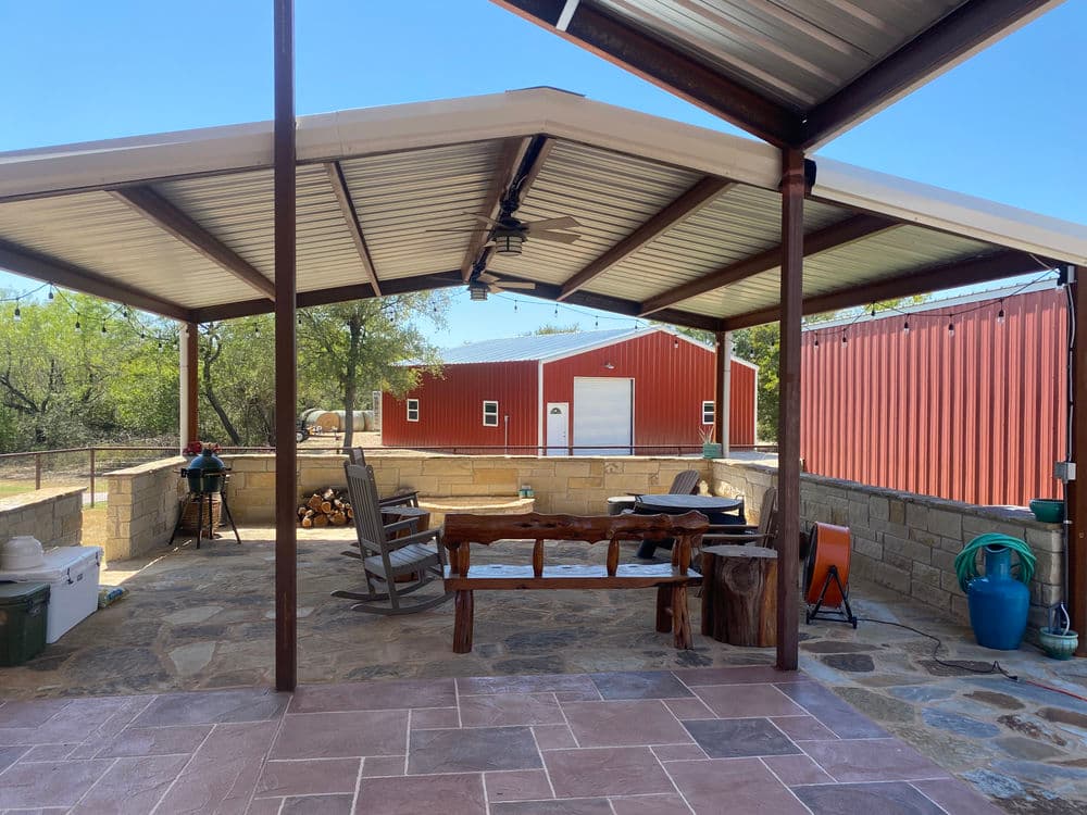 Outdoor patio area with shaded seating, stone fireplace, and red barn in the background.