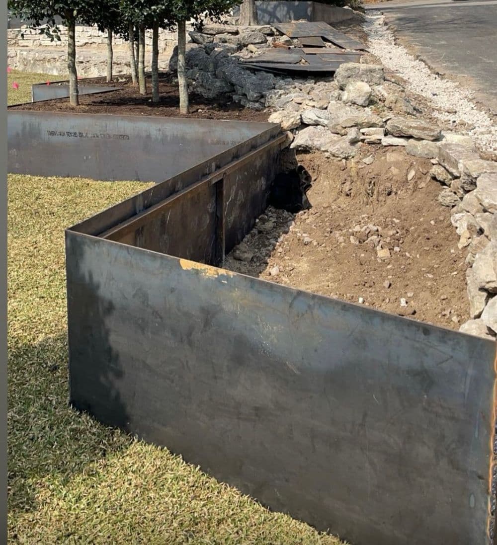 Metal garden bed under construction, surrounded by rocks and grass, near a paved path.