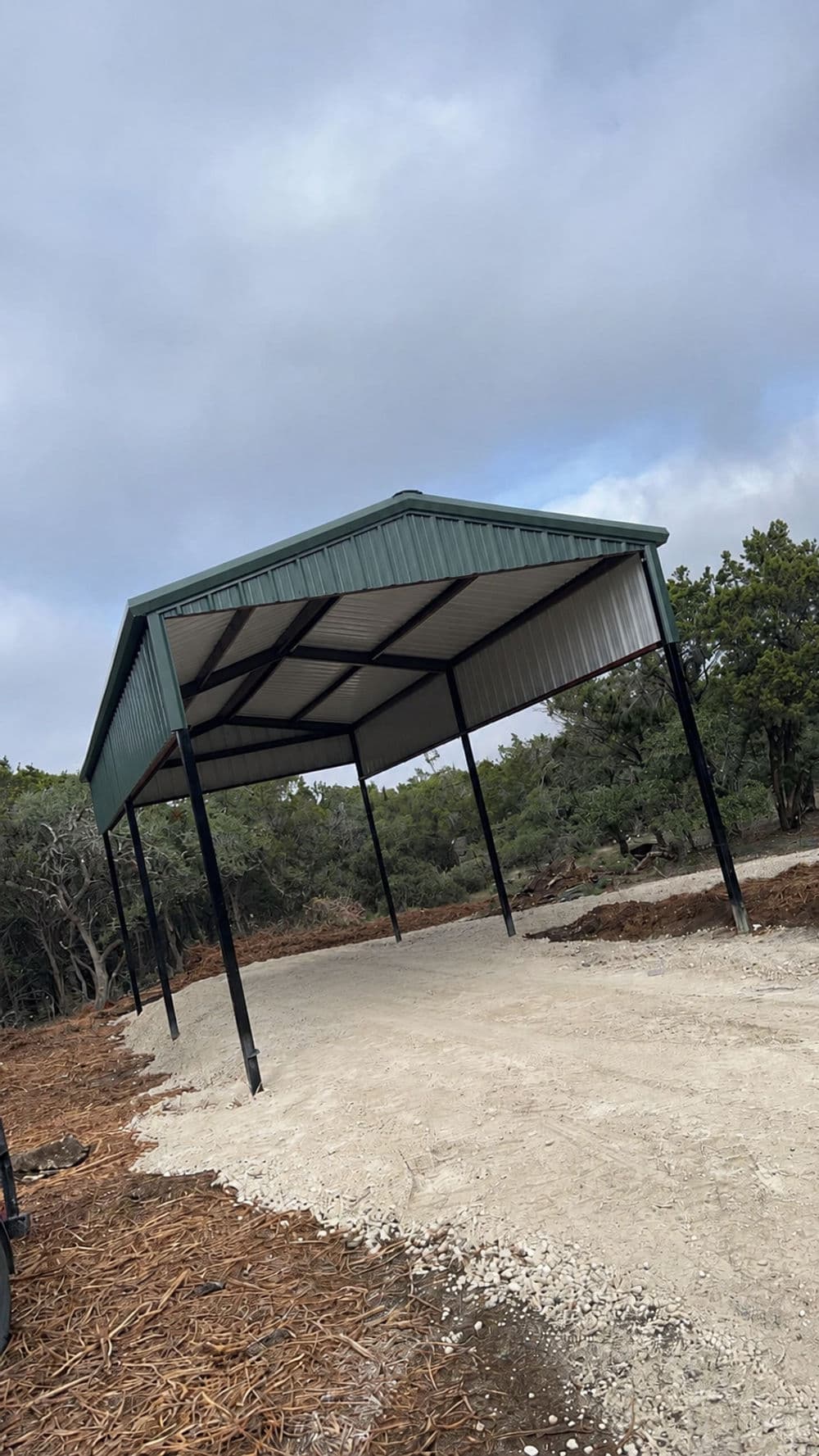 Metal carport structure on a gravel driveway surrounded by trees.