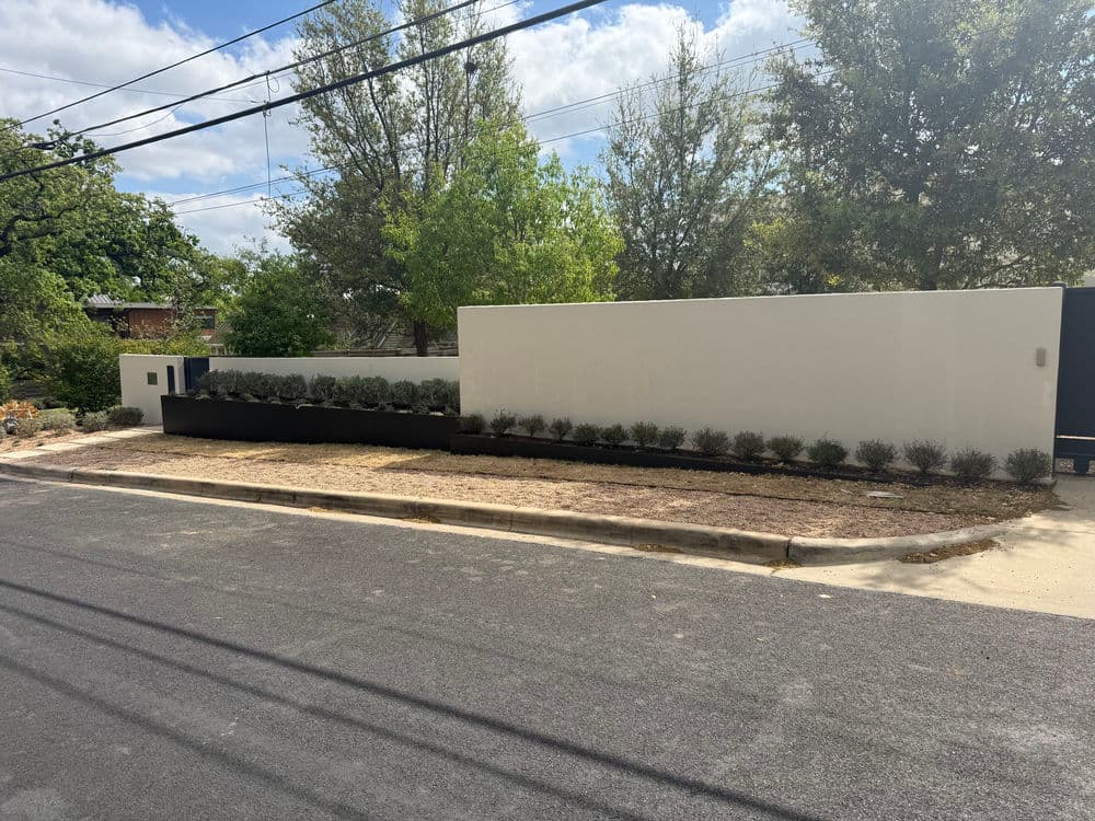 Modern white wall with landscaped garden, located on a quiet street with trees in background.
