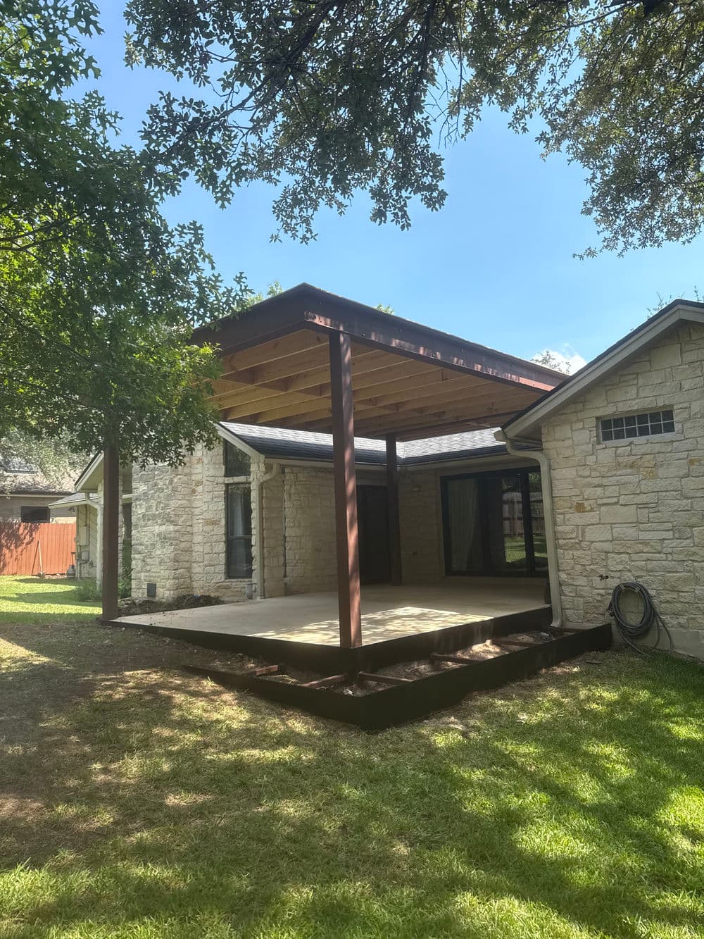 Backyard patio with wooden pergola, grass lawn, and stone house under a clear blue sky.