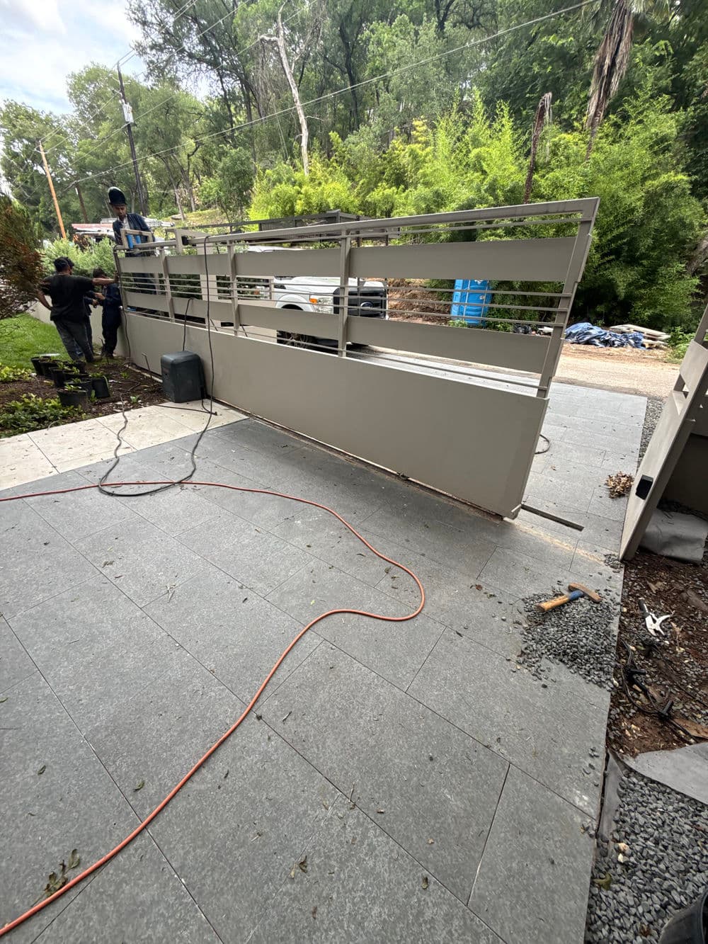 Metal driveway gate installation with workers and tools, surrounded by greenery.